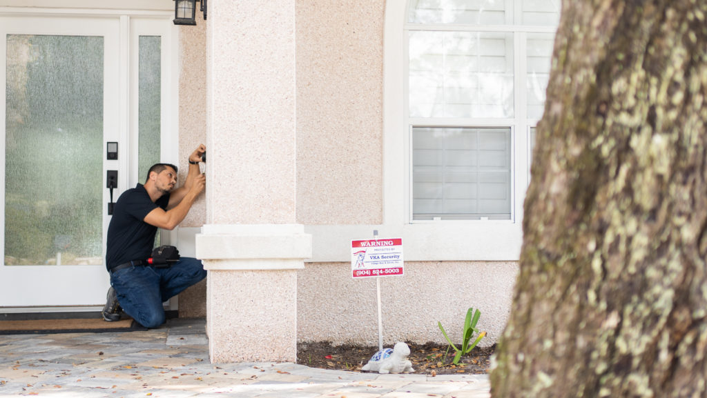 photo of an alarm tech installing an alarm system in front of the front door of a house with a VKA Security Alarm sign in front